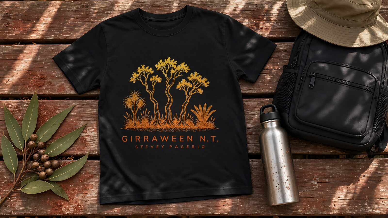Black kids t-shirt with the Girraween N.T. gum tree print in a yellow-to-orange fade, laid flat on a weathered timber bench with gum leaves, gumnuts, a steel water bottle, a black backpack and a bucket hat