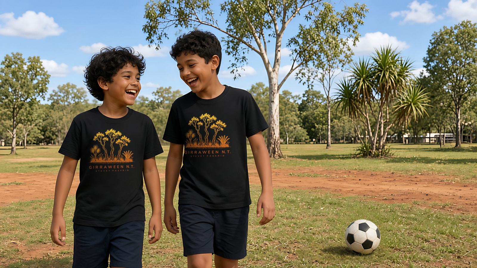Two laughing boys in black Girraween N.T. kids tees walking across a school oval next to a soccer ball, with pandanus and gum trees in the background